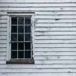 Peeling and cracking white paint on wood clapboard siding around an old double-hung window.
