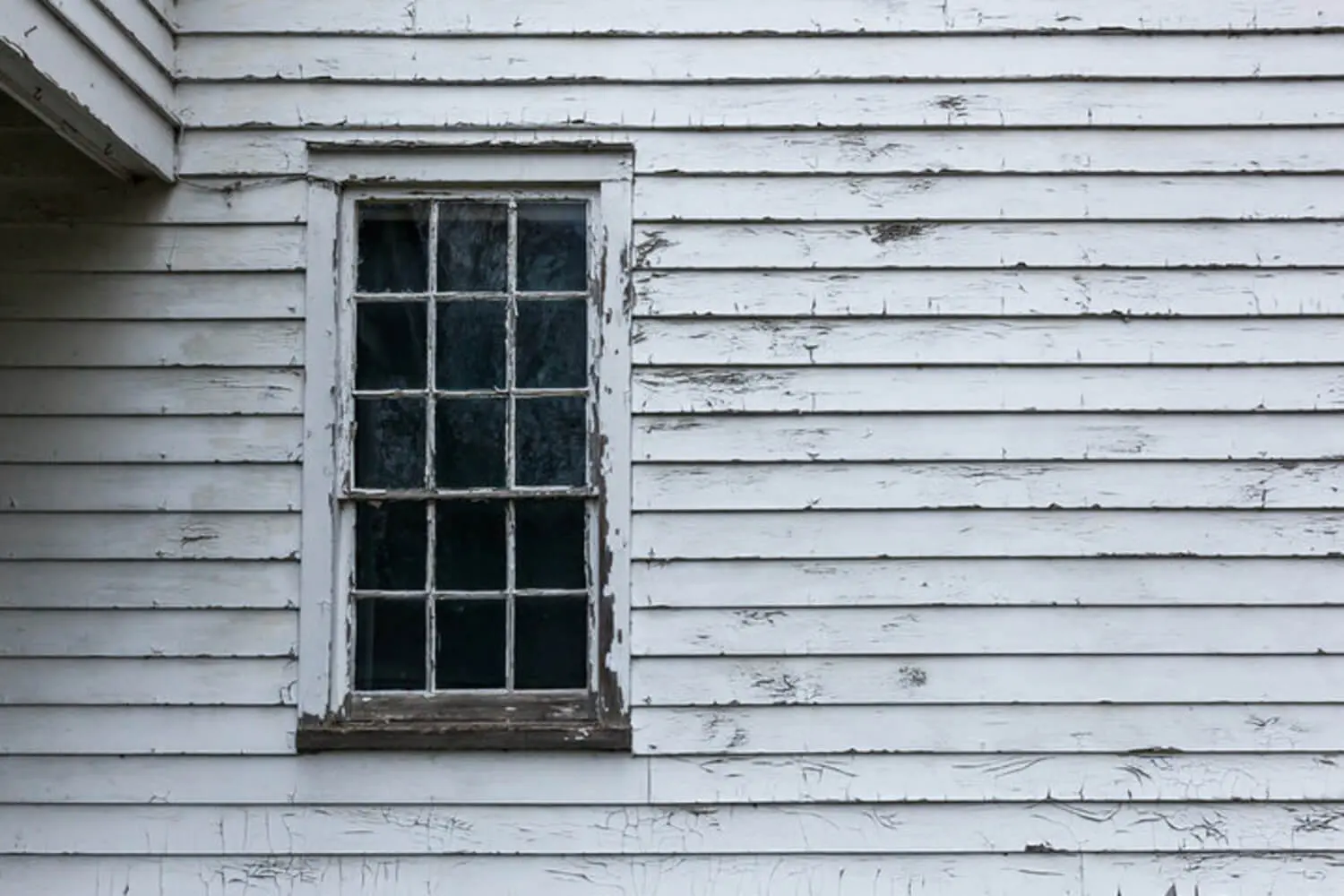 Peeling and cracking white paint on wood clapboard siding around an old double-hung window.