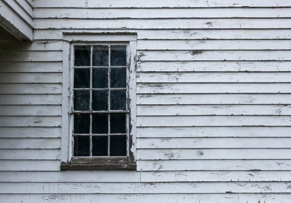 Peeling and cracking white paint on wood clapboard siding around an old double-hung window.
